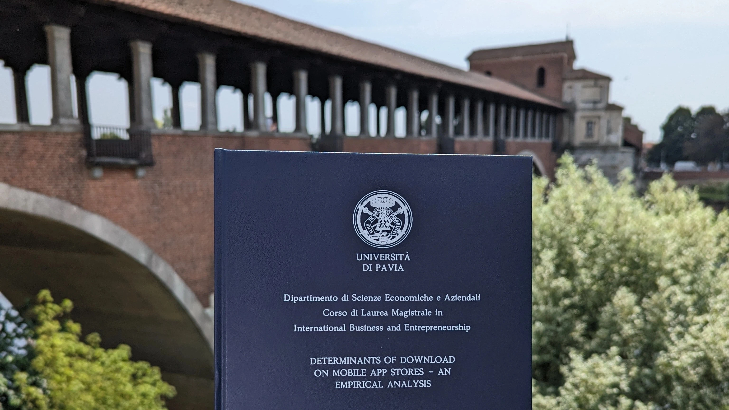 The bound copy of the thesis, a dark blue hardcover stamped in silver with the University of Pavia seal and the title 'Determinants of Download on Mobile App Stores – An Empirical Analysis', held up in front of Pavia's covered bridge over the Ticino on a sunny day.